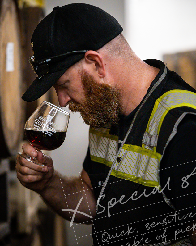 Bearded man in a cap and high-vis vest, a brewer, smells a glass of dark beer in a tasting session. 