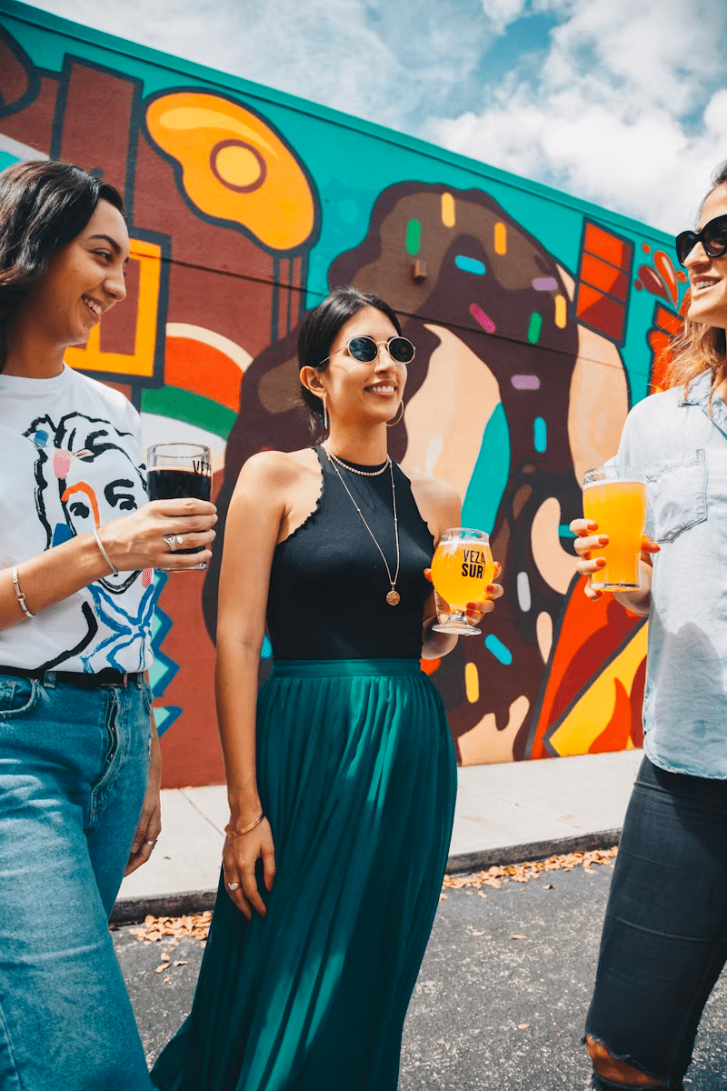 Three young women stand infront of a brightly coloured, painted wall. They are holding pints and smiling.