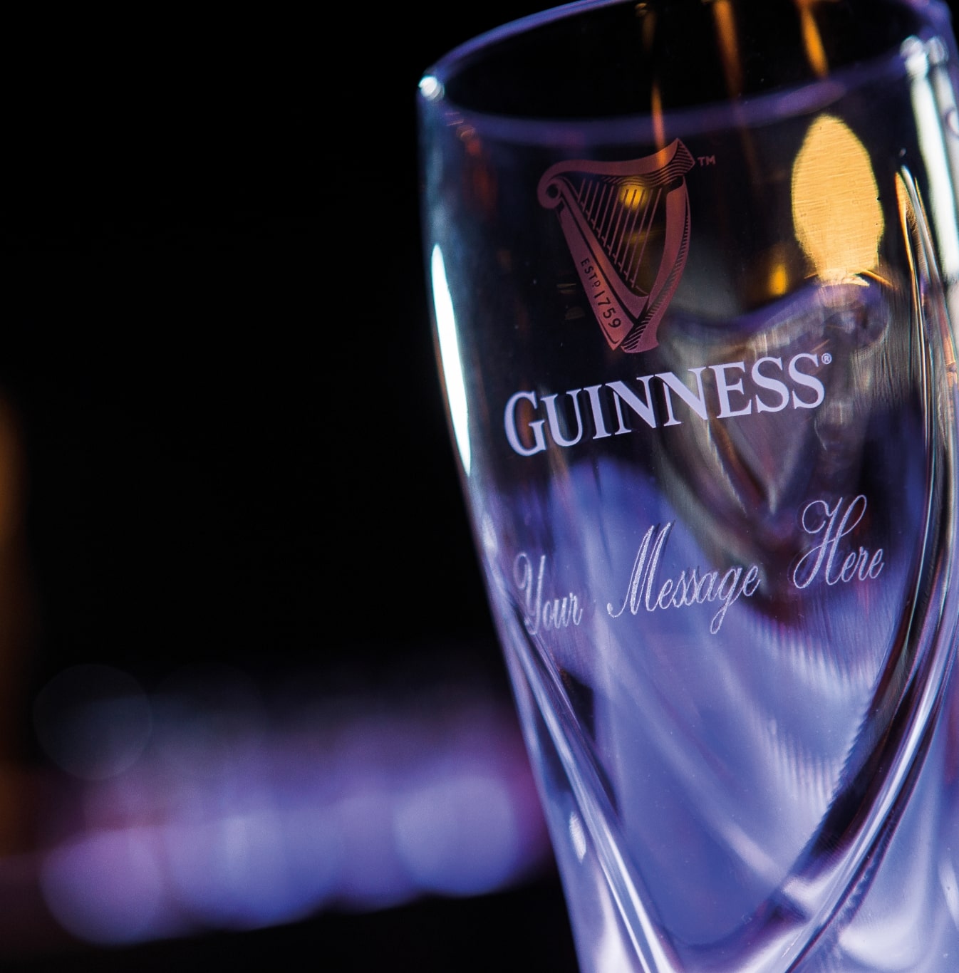 A close-up of a clear Guinness pint glass with the Guinness logo and "Your Message Here" engraved on it, against a dark, blurred background. 