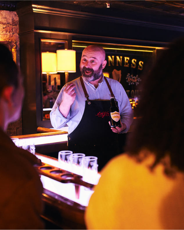 A man stands behind a bar holding a bottle of Guinness. He is talking animatedly to customers.