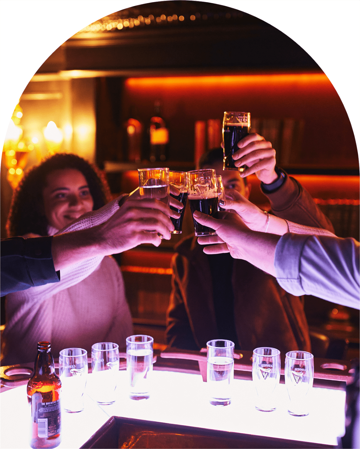 Several people raise dark beers in a toast, with a smiling woman in the background, and a light-up tray of small glasses on a bar counter. 