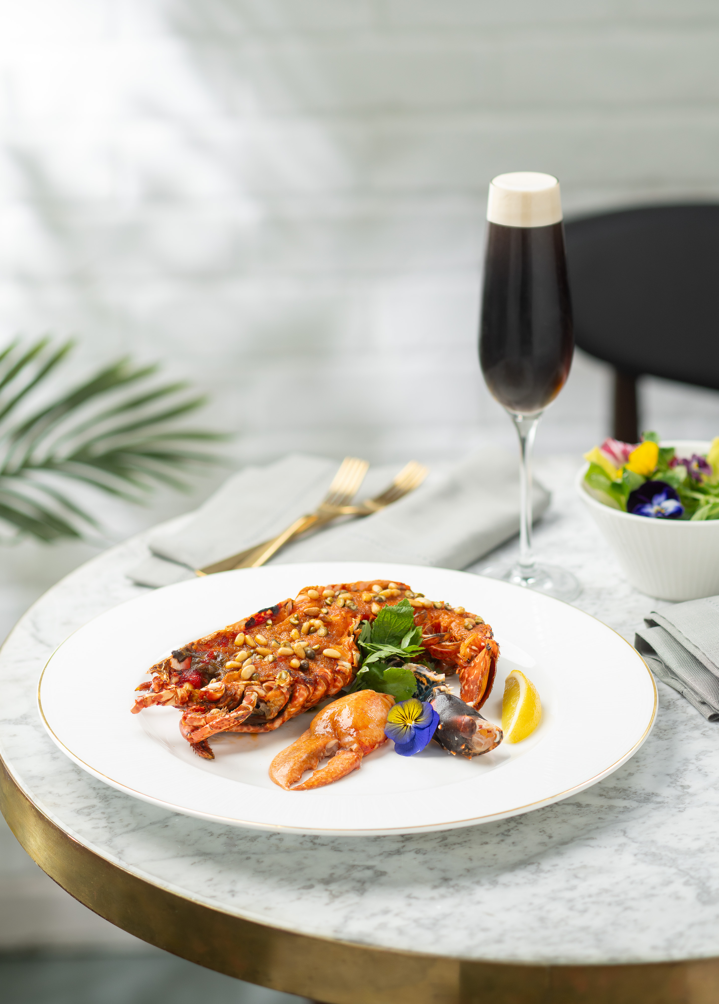 A white plate with cooked lobster, on a marble table. A tall glass with a dark beverage and a small salad bowl are in the background. 