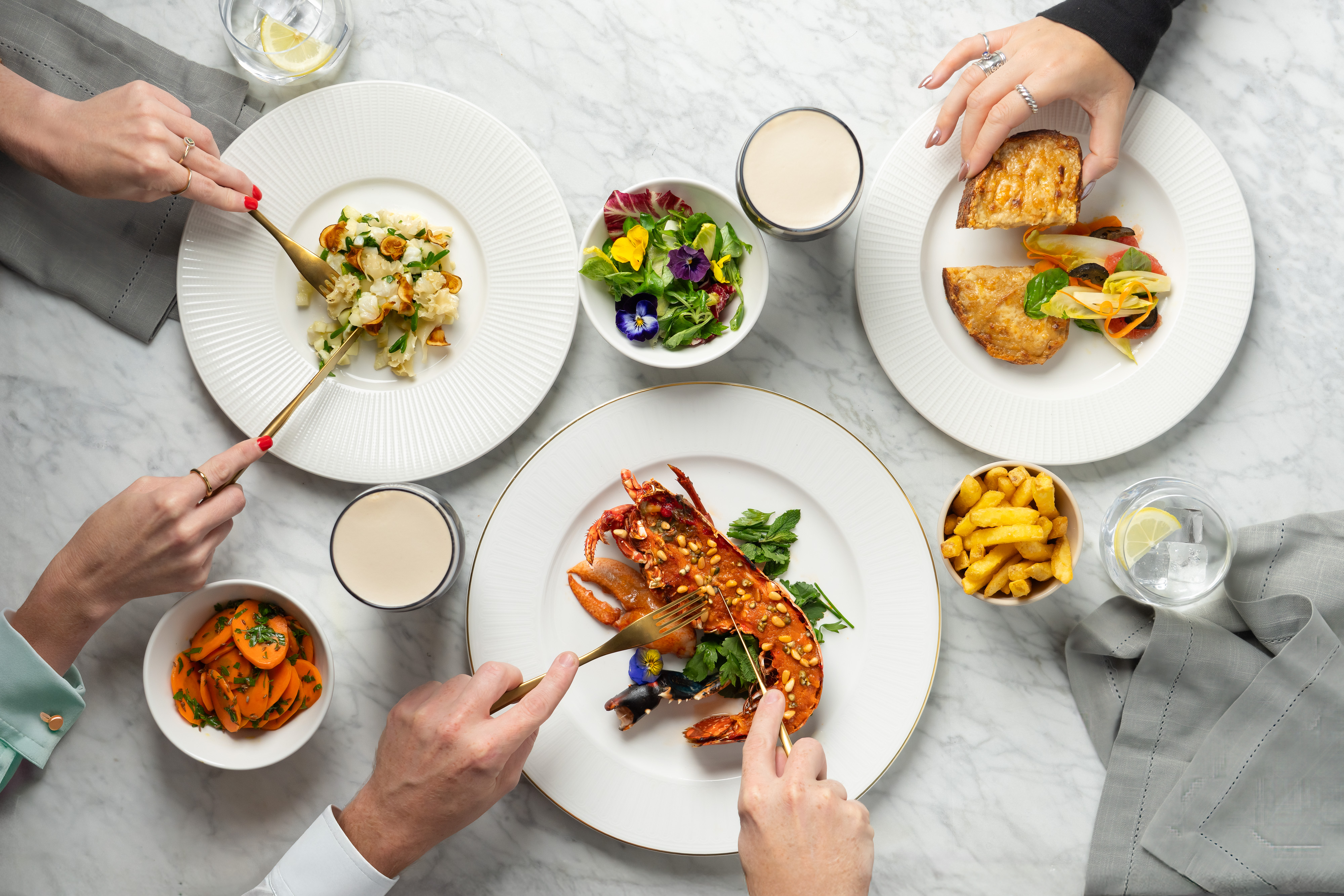 A top-down view of several people dining at a marble table, enjoying dishes like a whole lobster, cauliflower, pastries, salad, carrots, and fries.