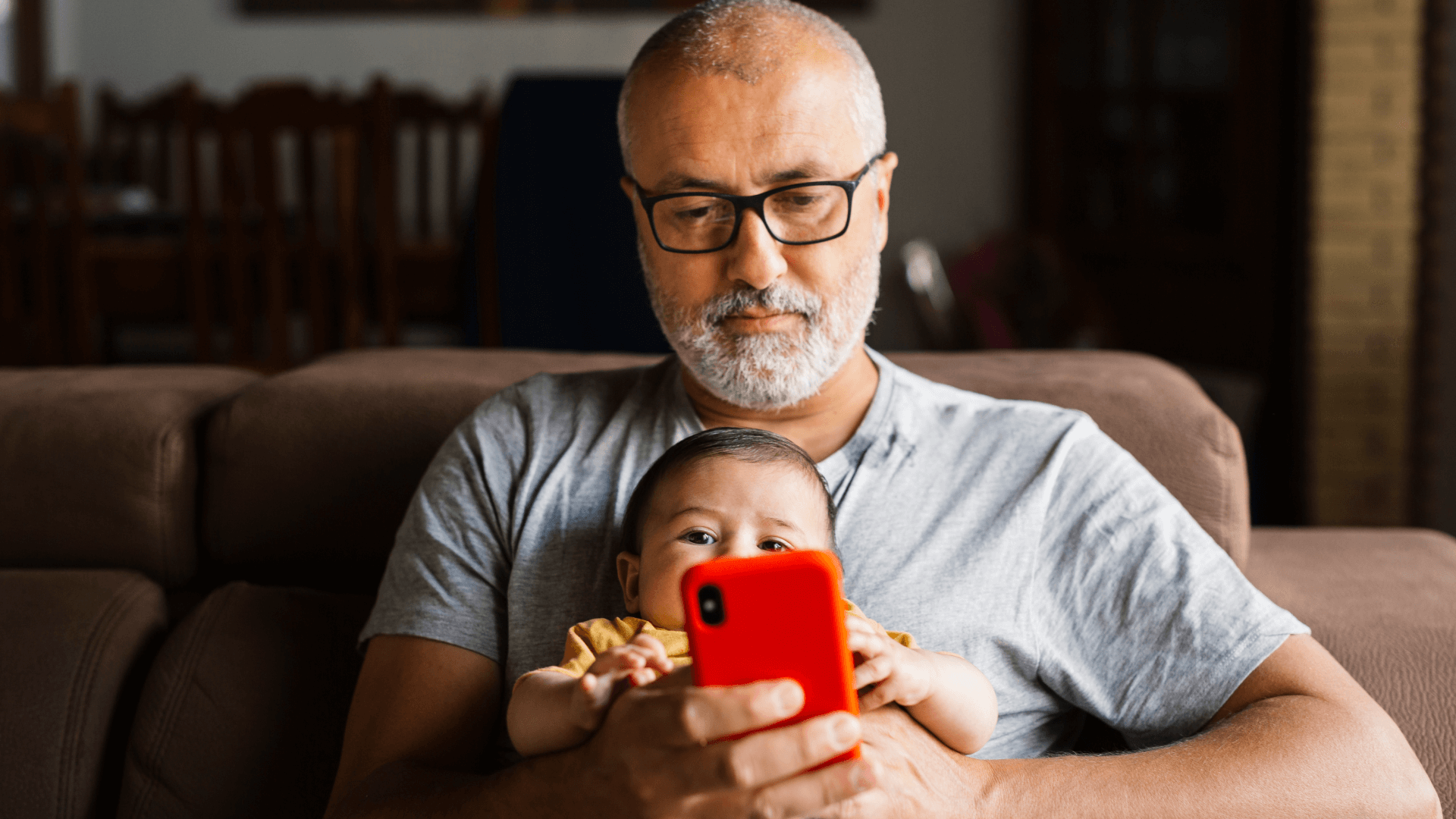 Image featuring a man with very short hair, glasses and a gray T-shirt, holding a baby while looking at this phone.