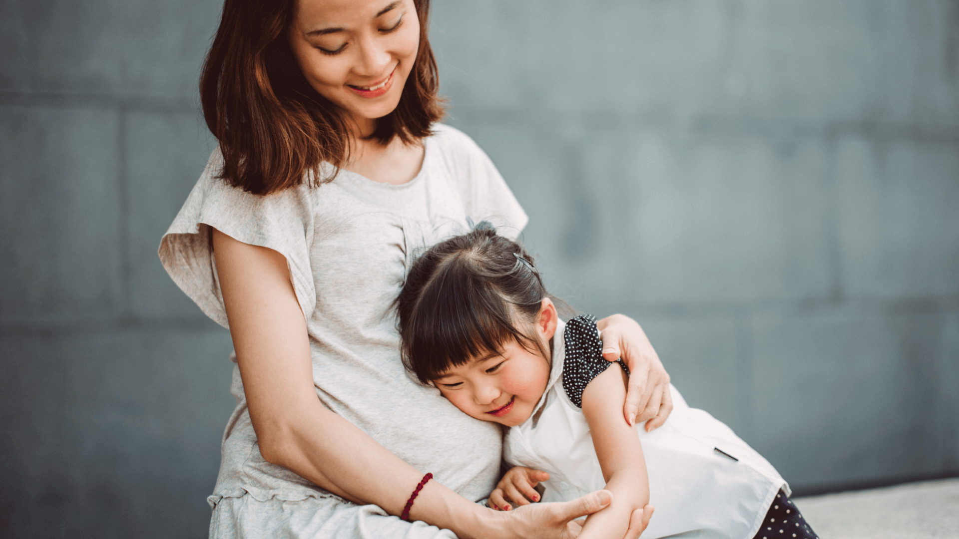 Image featuring a pregnant woman holding a young girl. Both of them are smiling.