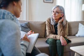 Woman in her 60s listens attentively sitting on a living room sofa while a woman on an opposite sofa take notes.