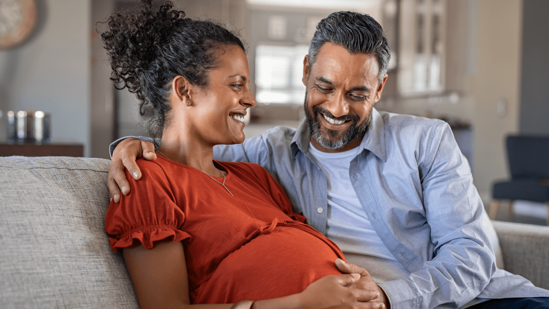 Happy father hands on expecting mother's baby bump while embracing her on couch