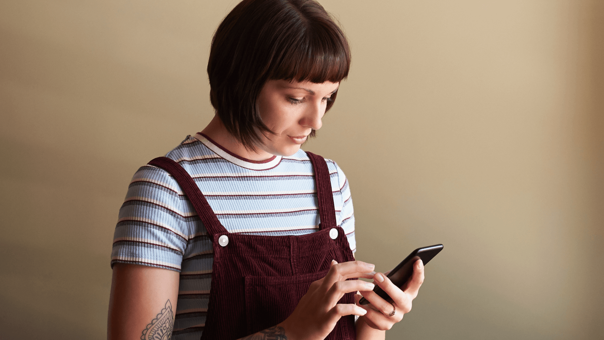 Image featuring a young woman with short hair, wearing a jumpsuit, looking at her phone.
