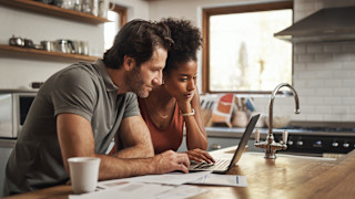 A man and a woman sitting together at a kitchen counter, reading from a laptop screen.