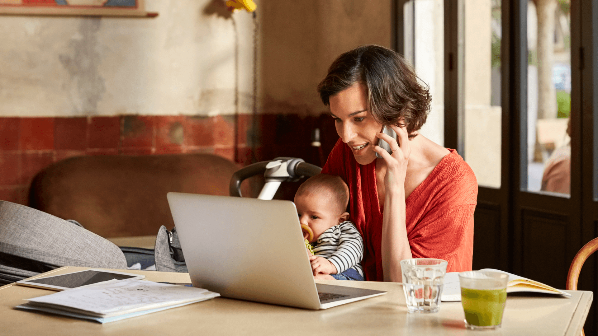 Image of a young woman with a baby in her lap talking on the phone and looking at a laptop.