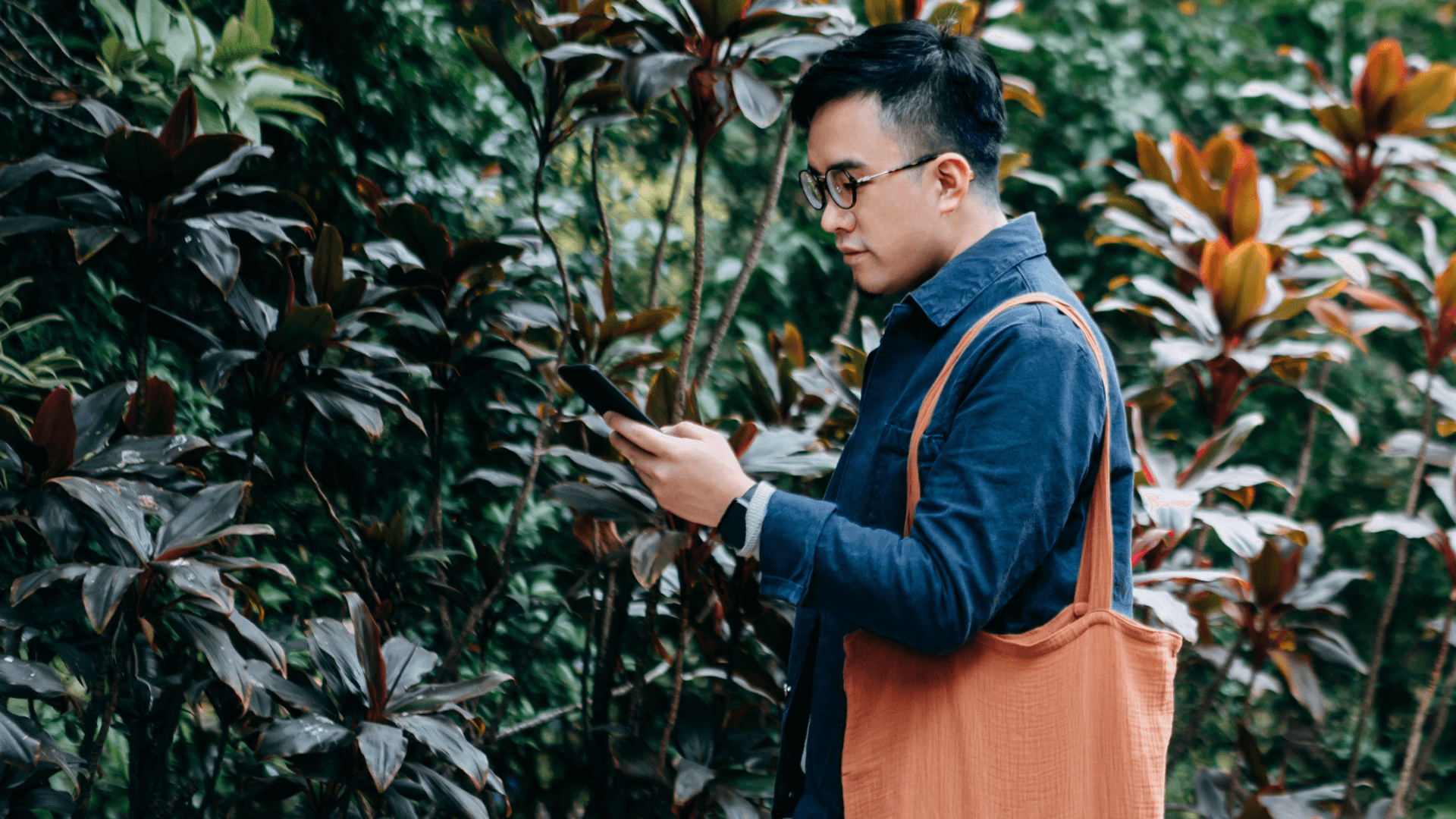 Image featuring a man wearing glasses, looking at his cellphone, with a lush garden in the background.