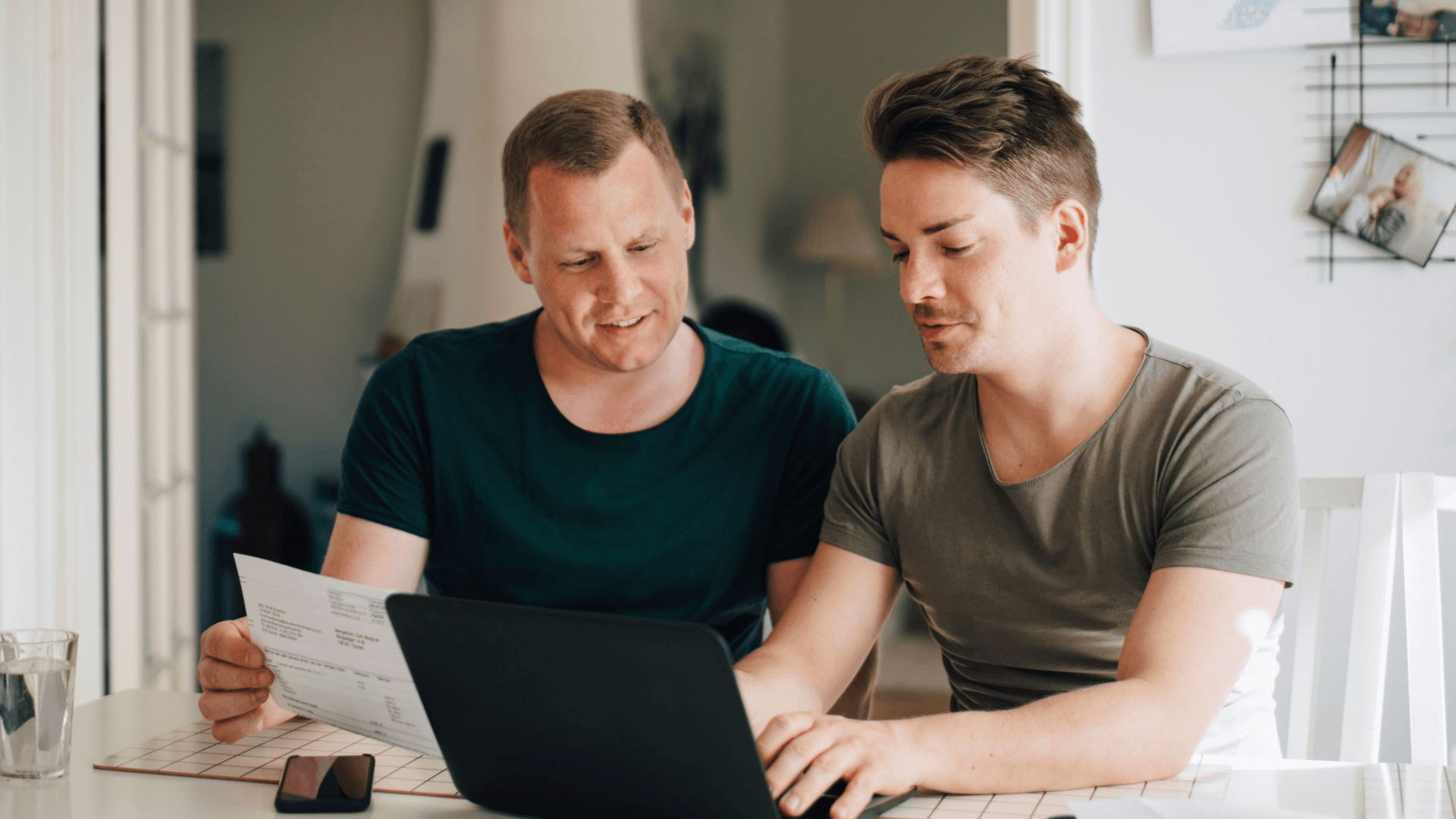 two men sit together in front a computer at home, one is typing and one is holding a bank statement