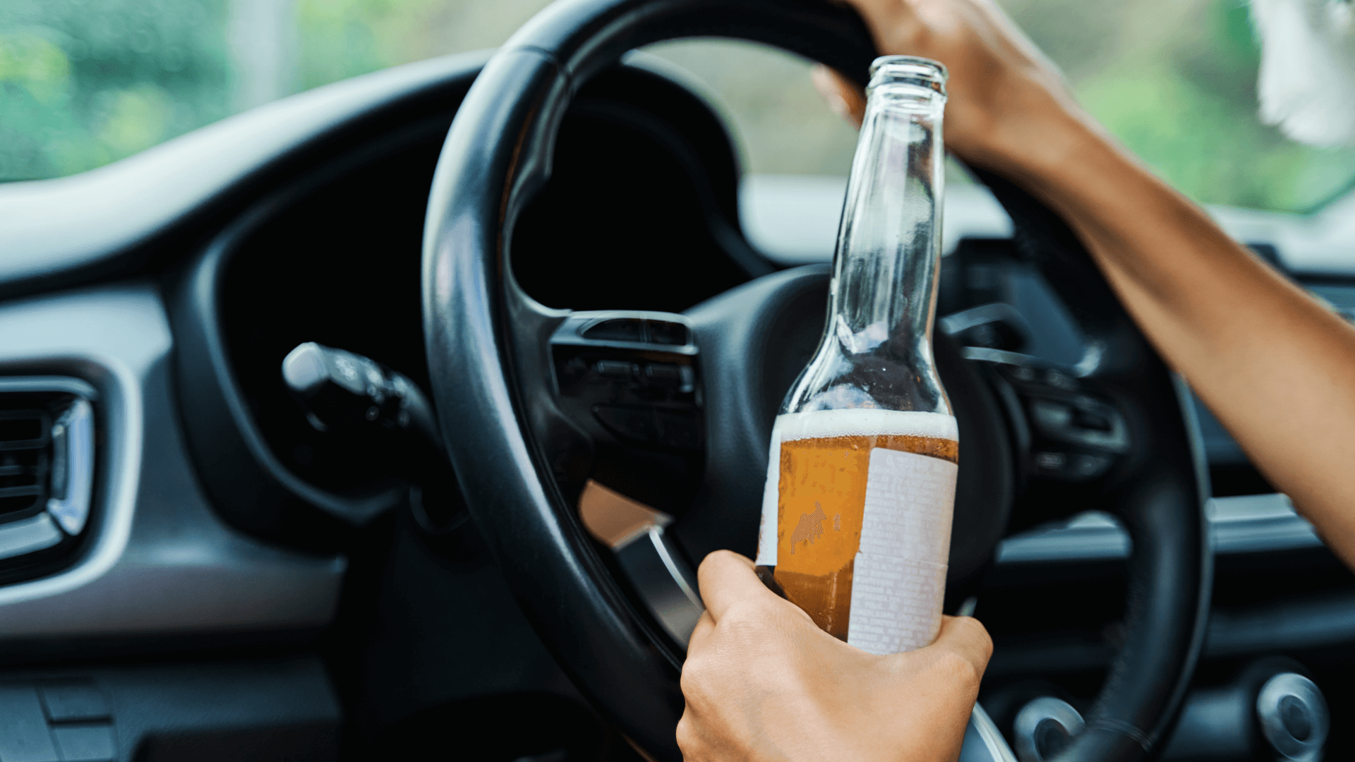 a woman's right hand grips a steering wheel while her left hand rests on it while holding a half-full bottle of beer