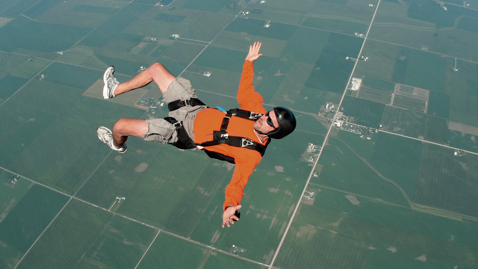 man in orange jumper skydiving over a large green expanse of fields and roads