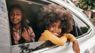 A young child sitting in the backseat a car with her arm hanging out the window, with another child sitting next to her