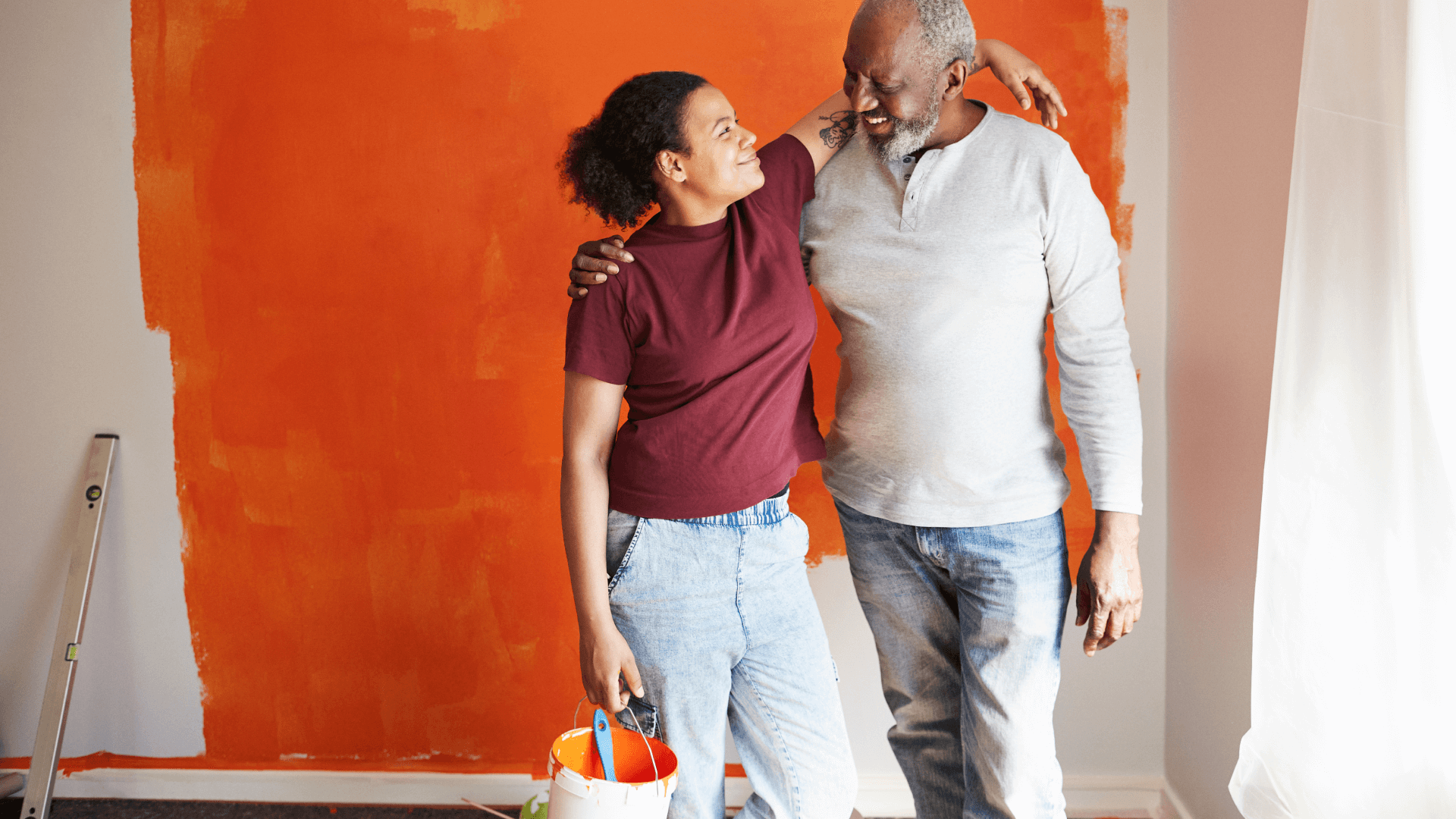 two African Americans, one an on older man and one a younger woman, stand in front of a newly-painted orange wall smiling with their arms around each other, the woman is holding a paint bucket and brush 