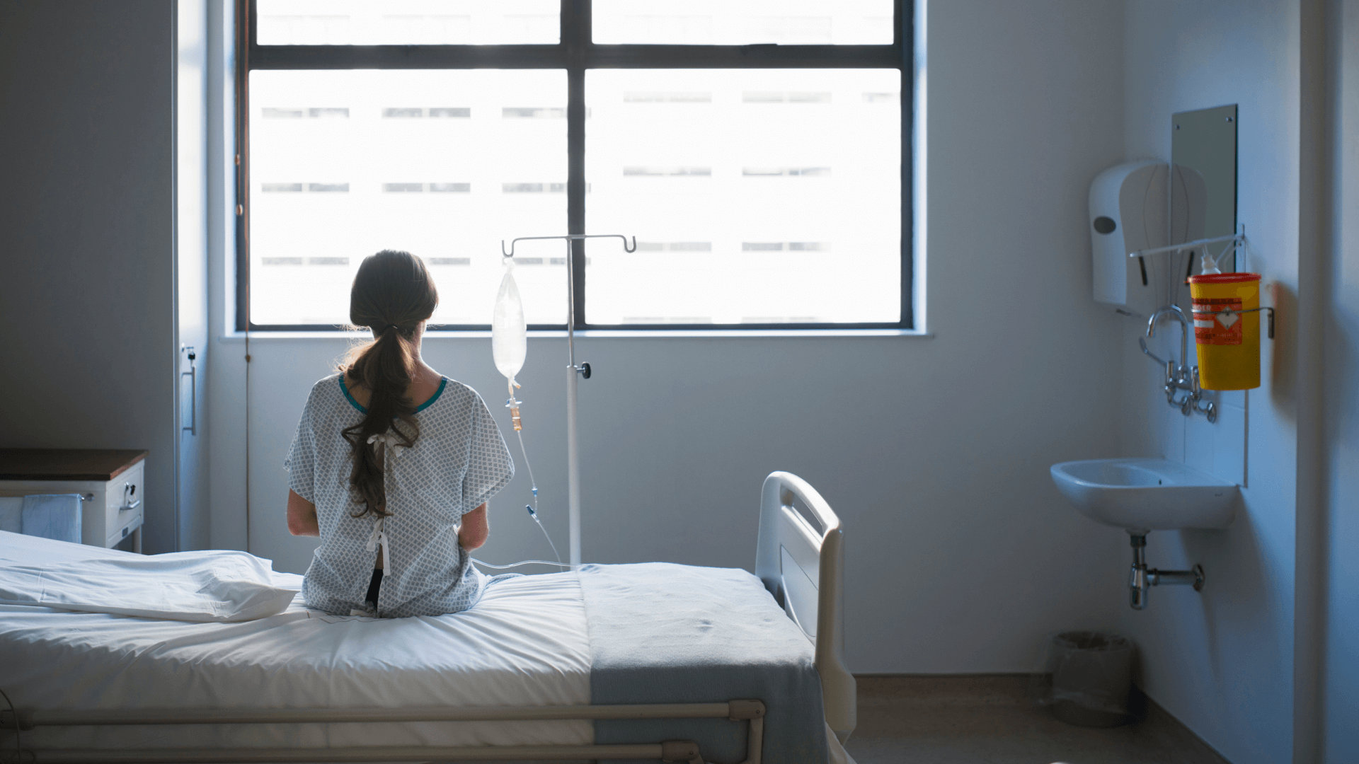 woman with long brown hair sits on a bed in a hospital room, she looks out a window and her back is to us, there is an IV pole next to her