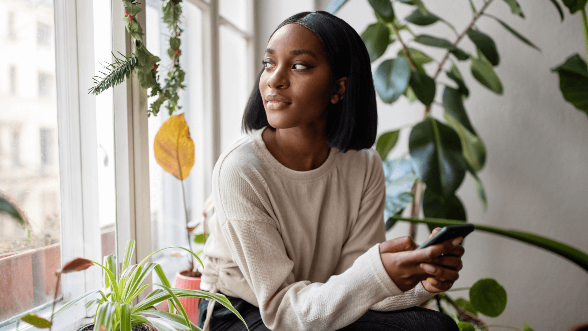 Woman sitting on the windowsill with mobile phone and looking away.