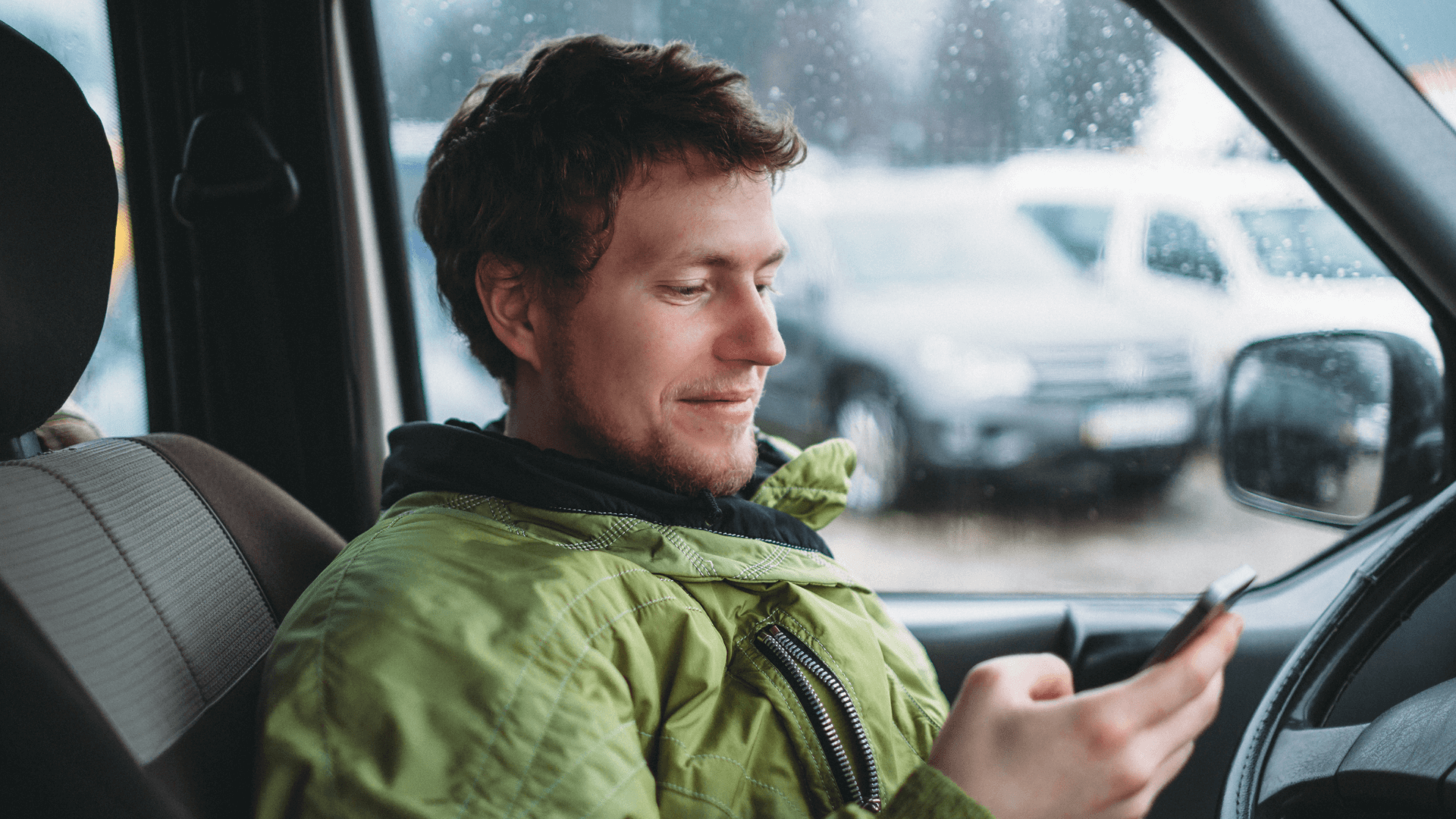 young man in green jacket sits at the wheel of a car and smiles while looking at his phone in the right hand