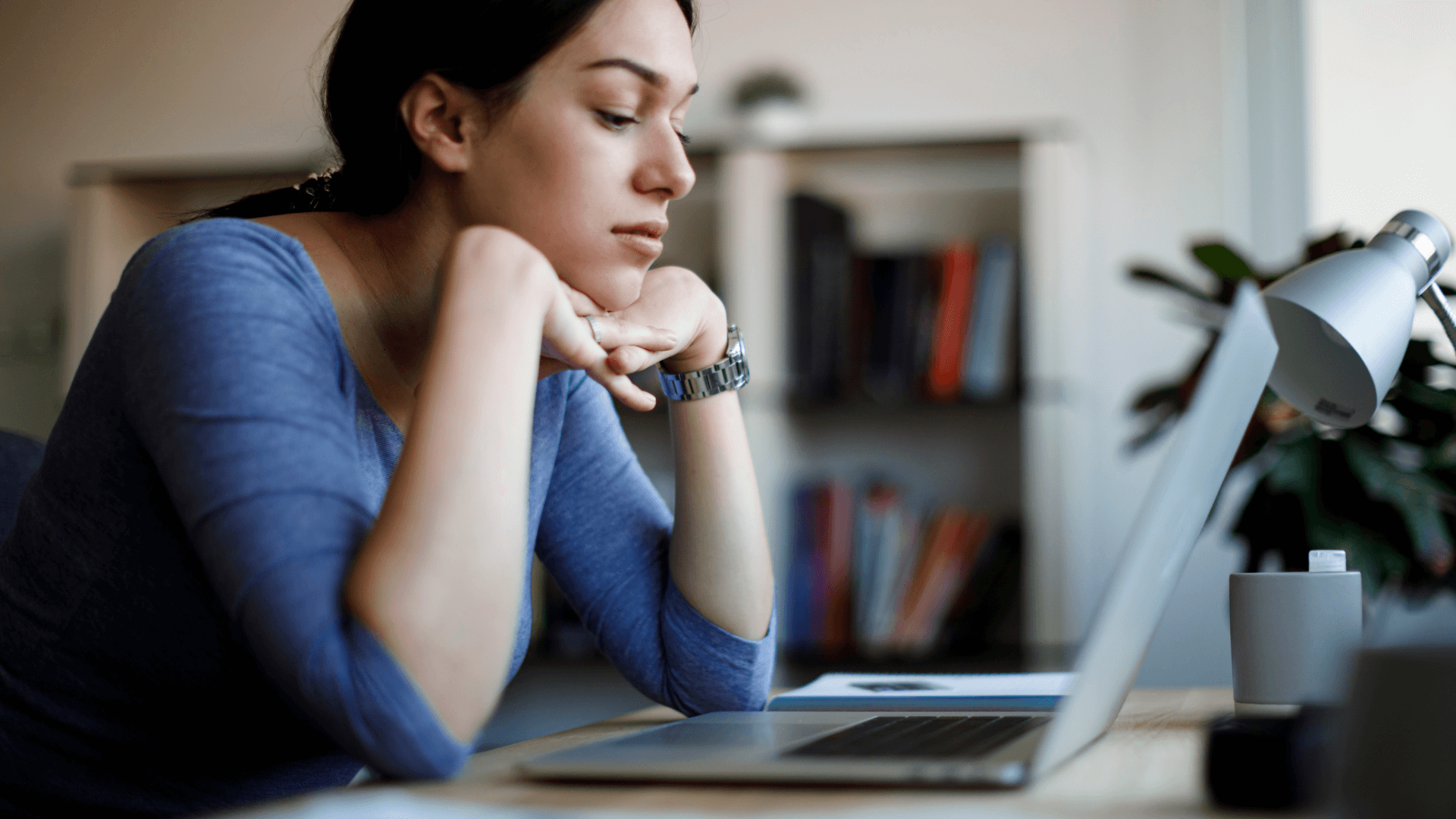 a woman in her 30s sits at a table in front of a laptop, her head rests on her hands as she stares at the screen