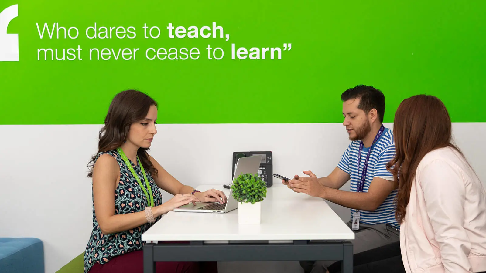 Group of people working on a desk by a wall with a quote: "Who dares to teach must never cease to learn"