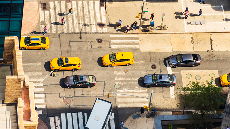 Overhead view of a busy city street with moving cars on the road and pedestrians walking on the sidewalk. 