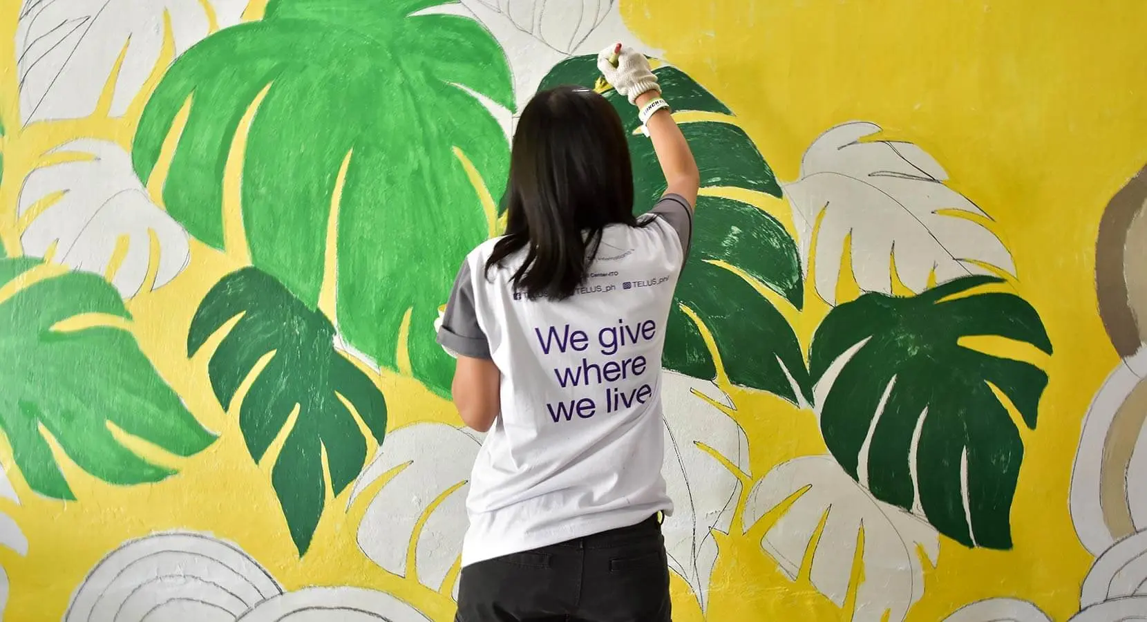 A woman wearing a "We give where we live" shirt painting a leaf wall mural