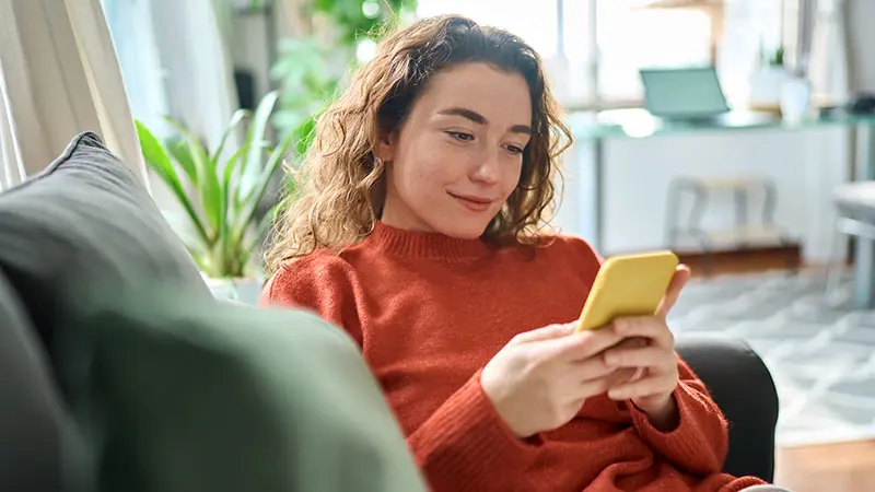 Woman sitting on a couch smiles as she looks at her mobile device. 