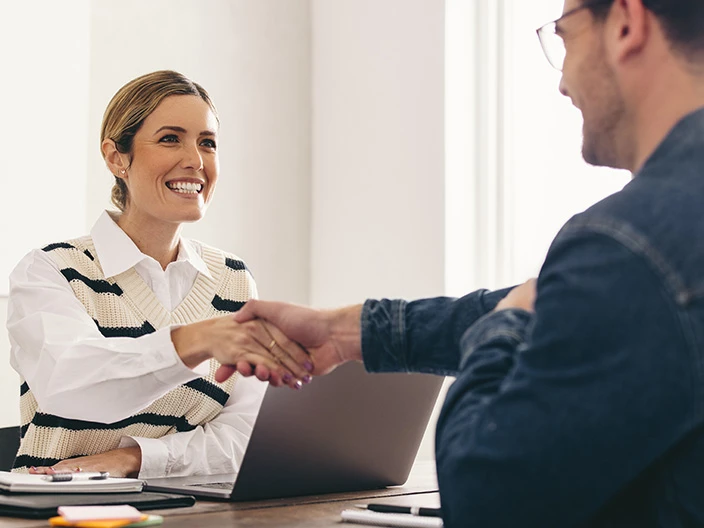 A female professional sitting at a desk shaking the hand of a male professional.