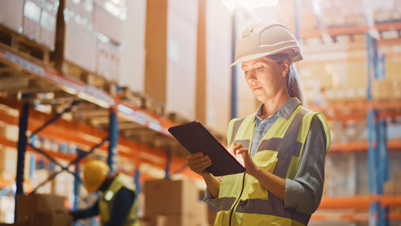 A woman warehouse worker wearing a hard hat looking at a tablet.