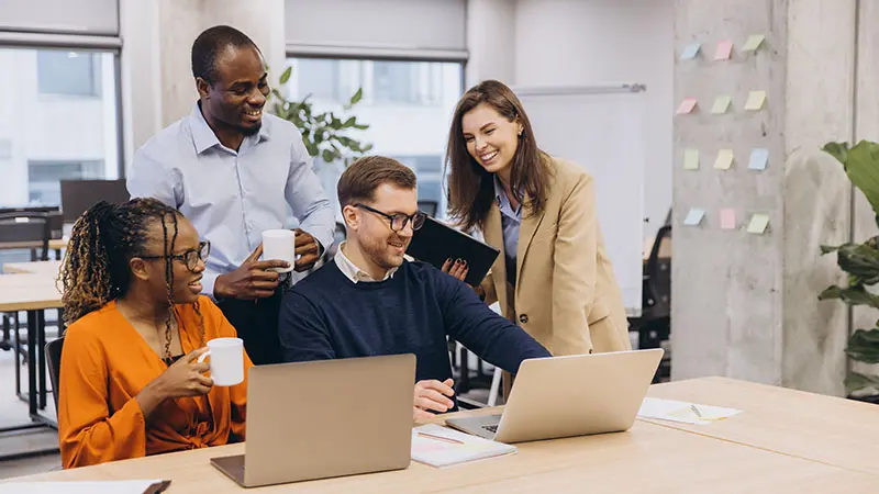 Four people working at a desk in an office, two are sitting in front of laptops and two are standing behind them.