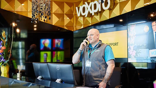 Man standing and talking on phone at a desk in front of a wall that says 'voxpro'