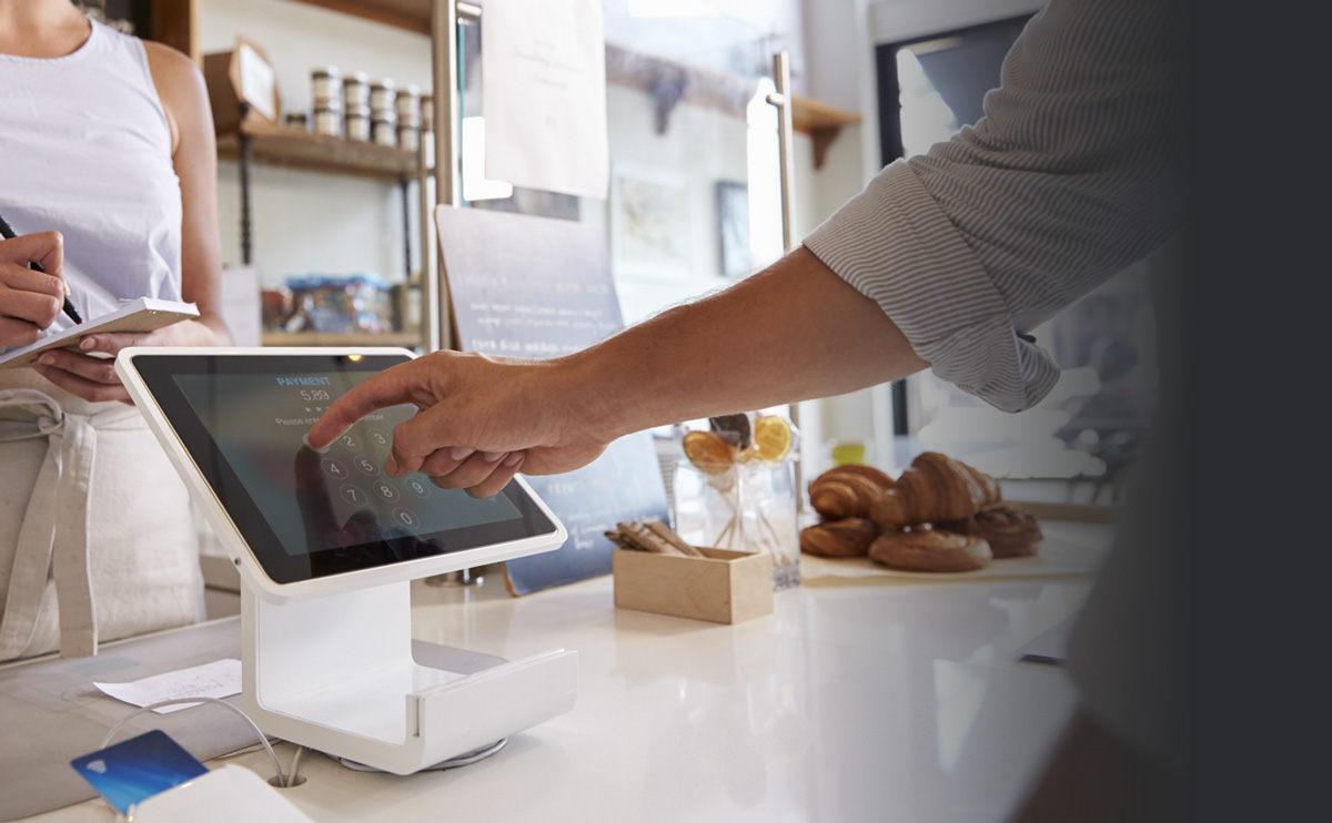 A woman taking an order at a coffee shop while the customer uses a touch-screen checkout.