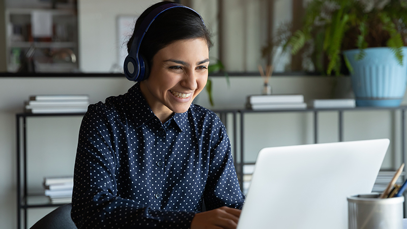 A young woman wearing headphones sitting at a laptop computer smiling.