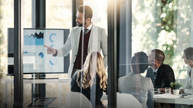 A male points to charts on a screen while his colleagues look on. 