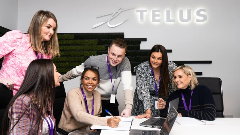 Photo of TELUS International team members in an office setting, gathered around laptop computers and taking written notes