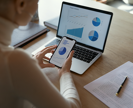 A woman looking at charts and graphs on a smartphone and a laptop.