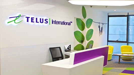 Desk in front of wall with TELUS International and a branch of a plant on it