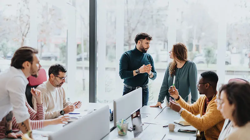 A group of people gathered around a boardroom table working on laptops and talking.