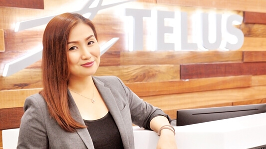 Woman in a suit leaning on a front desk smiling in front of a wood wall with a backlit TELUS sign