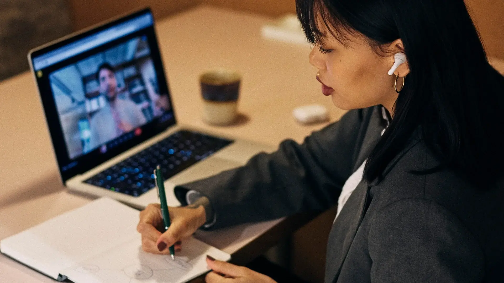 Image of a person writing notes with pen and paper and a laptop open to a meeting currently taking place