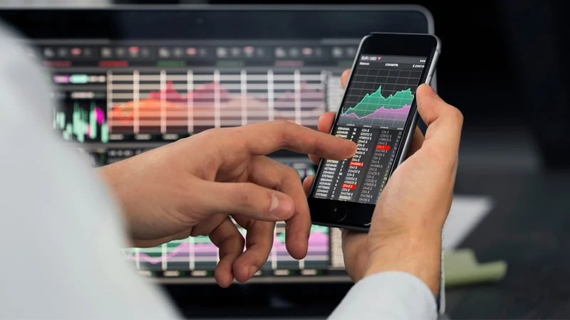 Person scrolling through financial information on mobile phone with laptop in the background