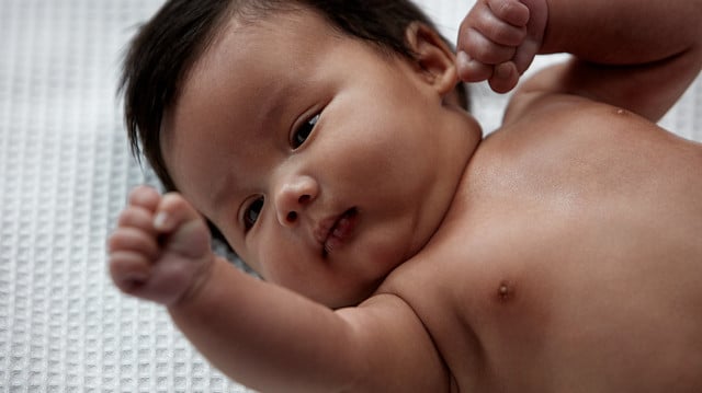 A baby lies on a textured white blanket, raising one hand and resting its other hand near its ear. The baby has short dark hair and is looking slightly to the side.