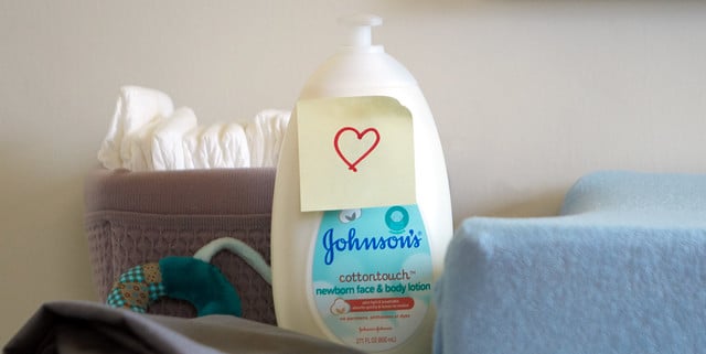 A bottle of Johnson’s newborn face and body lotion with a heart-drawn sticky note, placed beside a bin with towels, a pacifier, and blankets.