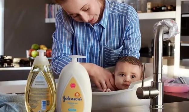A woman bathes a baby in a kitchen sink with two bottles of Johnson’s baby products nearby. Fruit and kitchen items are in the background in this well-lit kitchen.