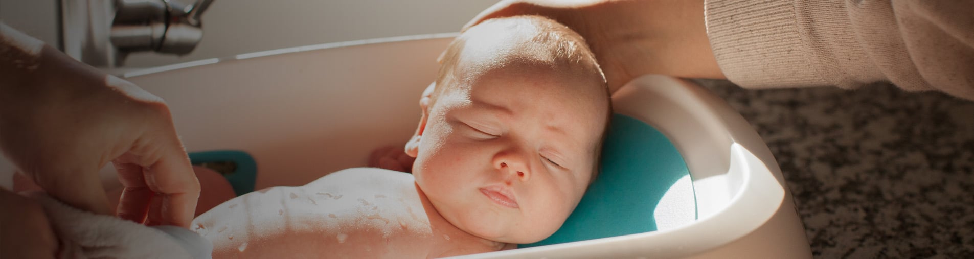 Baby bathing in a small tub, being gently washed and supported by adult hands, situated in a sunlit bathroom with a granite countertop.