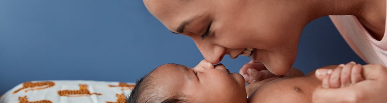 Mother touching noses with her baby, both smiling, with a blue background and baby blanket with animal prints.