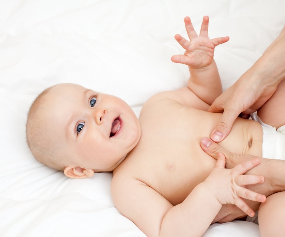 Smiling baby lying on a white bed while an parent's hands gently touch its belly.