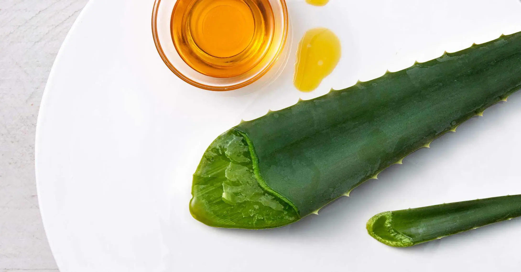 Aloe vera leaf and a small bowl of orange liquid on a white plate, representing natural ingredients.