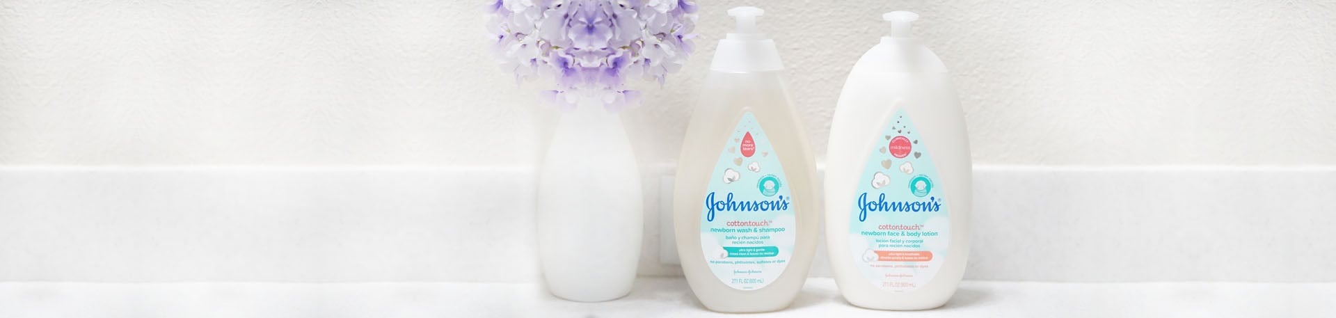 Two Johnson's baby cottontouch lotion bottles are placed on a white surface next to a vase of purple flowers.